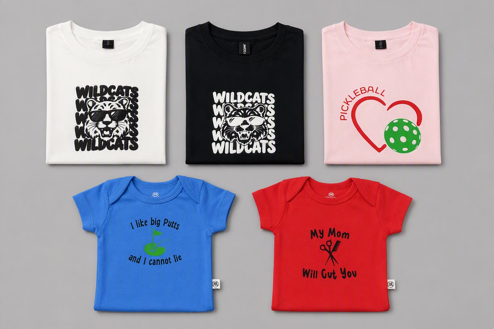 Five of Heather's custom pieces laid flat: two Wildcats tees, a pickleball heart tee and two baby onesies